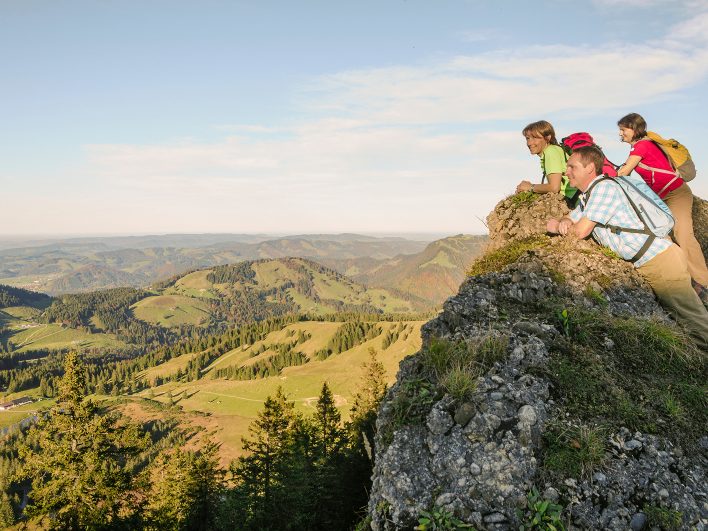 Drei Wanderer lehnen an einem Felsvorsprung und genießen die weite Aussicht über grüne Almen, Fichtenwälder und sanfte Hügel im Bregenzerwald.