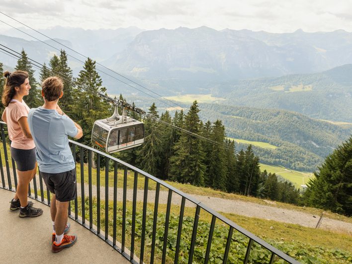 Paar an einer Aussichtsplattform an der Bergstation der Seilbahn Bezau, eine Gondel schwebt über den Fichten ins Tal, dahinter die Berge des Bregenzerwalds.