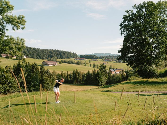 Golferin beim Abschlag auf einem grünen Golfplatz im Bregenzerwald, umgeben von alten Bäumen, Wiesen und Bauernhöfen im Abendlicht.