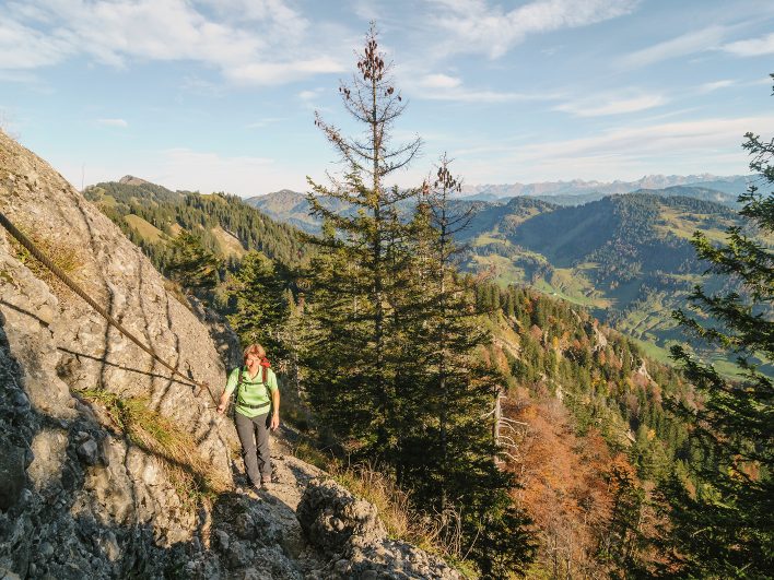Wanderin hält sich an einem Stahlseil fest und steigt einen felsigen Pfad an der Nagelfluhkette hinauf, rechts öffnet sich der Blick über herbstliche Wälder bis zu den Alpengipfeln.
