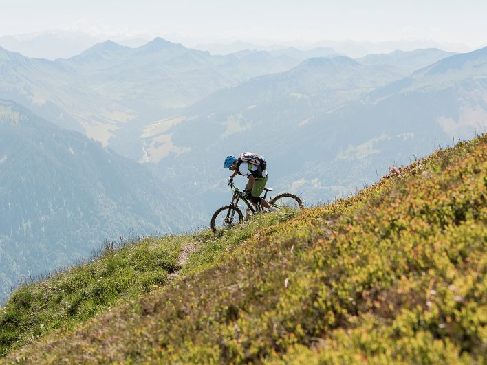 Mountainbiker fährt einen schmalen Trail entlang eines steilen Grashangs, dahinter reihen sich die Bergketten des Bregenzerwalds im Dunst.