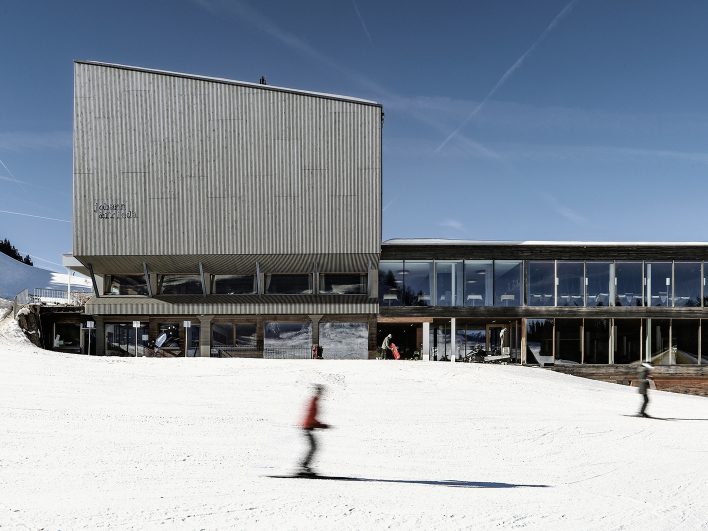 Restaurant Johann & Frieda am Almhotel Hochhäderich mit Holzfassade und großen Glasfronten, davor gleiten Skifahrer über die sonnige Piste der Alpenarena.