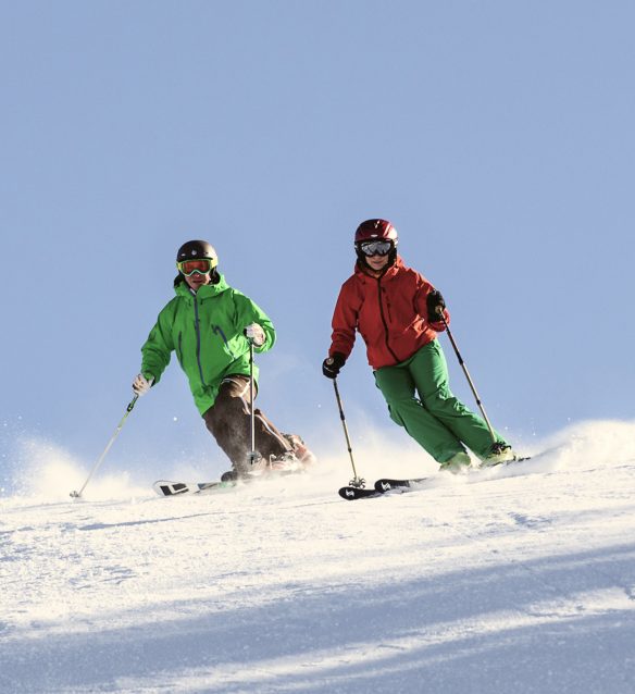 Zwei Skifahrer in grüner und roter Jacke carven nebeneinander durch den Pulverschnee, der bei jedem Schwung aufstiebt.