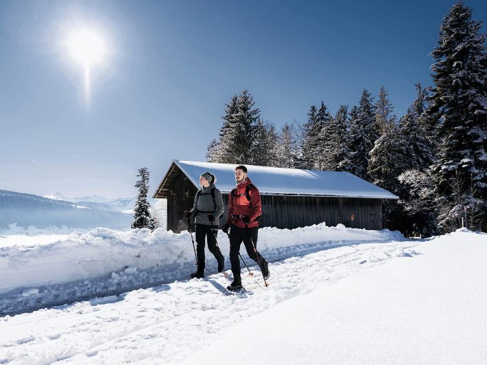 Paar mit Wanderstöcken auf einem geräumten Winterwanderweg im Bregenzerwald, vorbei an einem verschneiten Holzstadel in der Wintersonne.