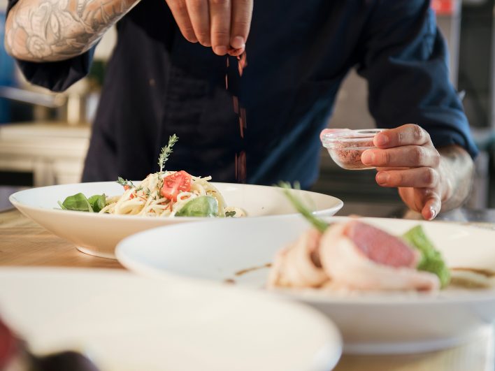 Koch bestreut beim Anrichten einen Teller mit Nudeln, Tomate und Thymian, daneben ein Teller mit rosa gebratenem Fleisch.