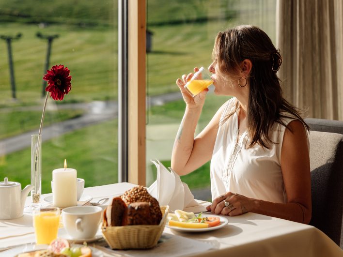 Frau frühstückt am Panoramafenster mit Blick auf die Wiesen und den Skilift und trinkt ein Glas Orangensaft.