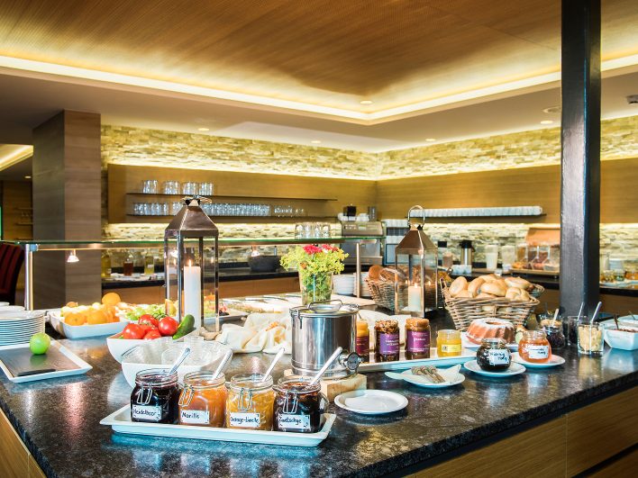 Frühstücksbuffet mit Marmeladen in Gläsern, Honig, Obst, Gemüse, Brotkörben und Gugelhupf auf einer Steintheke.