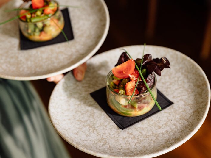 Serviceperson trägt zwei Teller mit Salat im Glas, garniert mit Tomate, Schnittlauch und rotem Blattsalat.