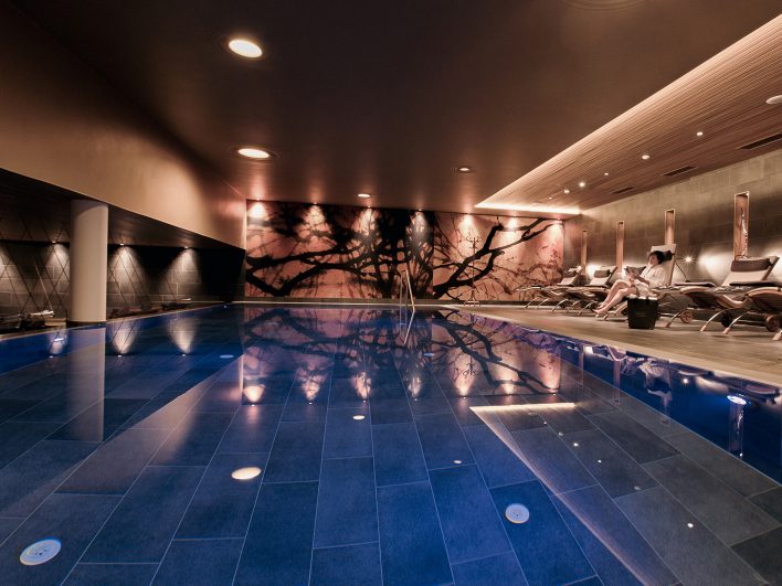Beleuchtetes Schwimmbecken im AlmSpa mit Wandbild aus Ästen, an der Seite Liegen, auf denen ein Gast liest.