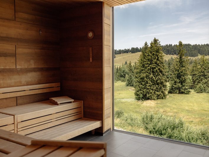 Leere Holzsauna im AlmSpa mit raumhohem Panoramafenster, draußen Fichten, Almwiesen und bewaldete Hügel im Sommer.
