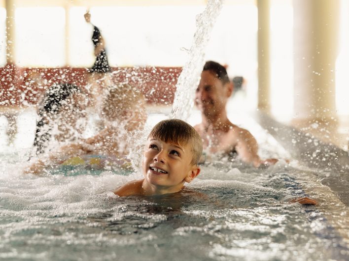 Junge schaut lachend zum Wasserstrahl im Panoramahallenbad hinauf, dahinter planschen Eltern und Geschwister.