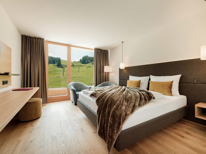 Doppelzimmer Hochhäderich mit Boxspringbett, Fellplaid und Sessel, bodentiefes Fenster mit Blick auf Almwiese und Skilift.