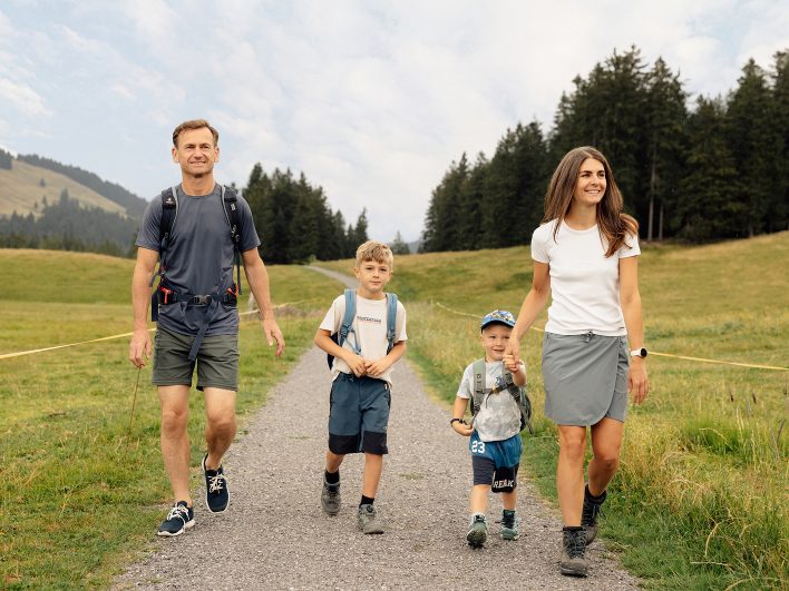 Eltern und zwei Kinder mit Rucksäcken wandern lächelnd auf einem Kiesweg über die Almwiese, die Mutter hält den Kleinen an der Hand.