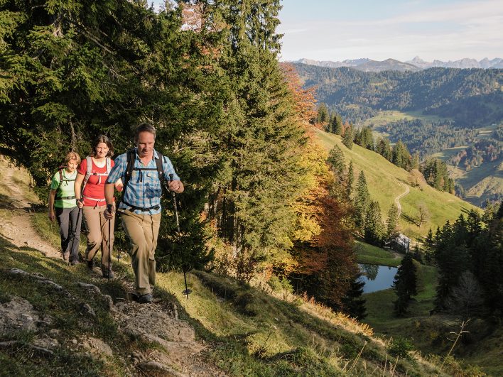 Drei Wanderer mit Stöcken auf einem schmalen Bergpfad im Bregenzerwald, darunter ein kleiner Bergsee, bunte Bäume und Almwiesen vor einem weiten Bergpanorama.