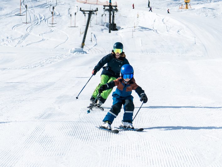 Ein Kind mit Helm und Skibrille fährt vor einer erwachsenen Begleitung die frisch präparierte Piste hinunter, im Hintergrund ein Schlepplift der Alpenarena Hochhäderich.