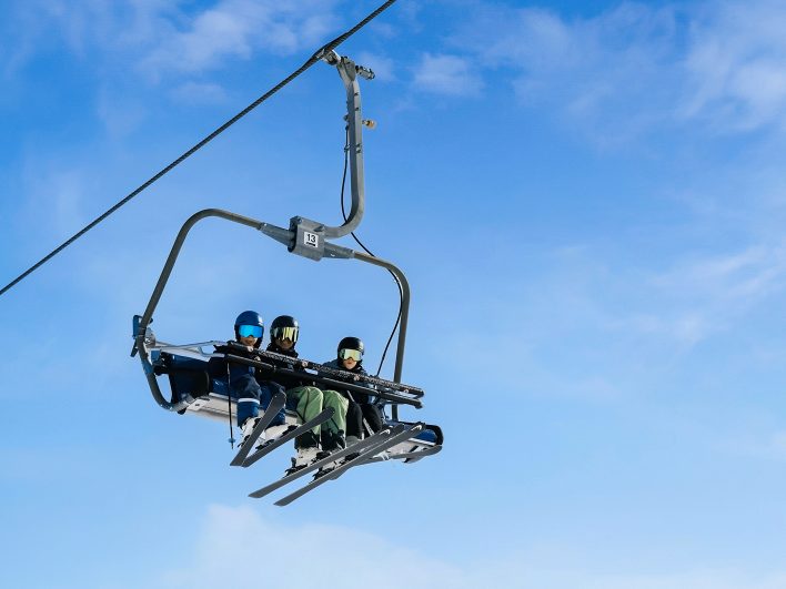 Drei Skifahrer mit Helm und Skibrille im kindergesicherten Vierersessellift der Alpenarena Hochhäderich vor blauem Himmel.