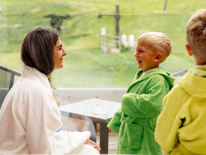 Zwei Jungen in grünen Kinderbademänteln lachen mit ihrer Mutter im weißen Bademantel auf dem Balkon mit Blick zur Wiese.