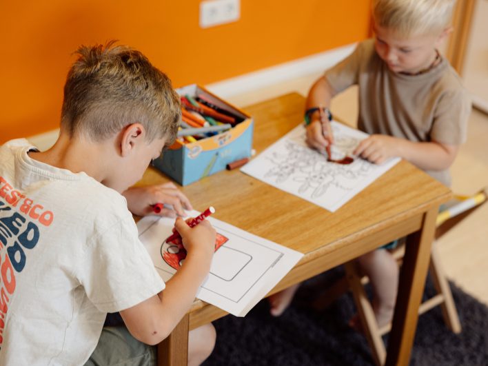Zwei Jungen malen konzentriert Ausmalbilder mit bunten Filzstiften am Kindertisch vor einer orangefarbenen Wand.
