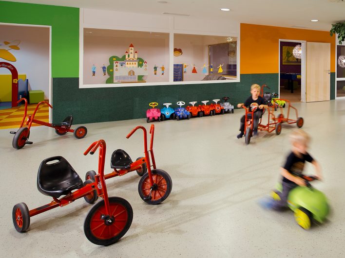 Zwei Kinder flitzen mit Dreirad und Rutschauto durch die Indoor-Kinderwelten des Almhotels Hochhäderich, an der Wand parken bunte Bobbycars.