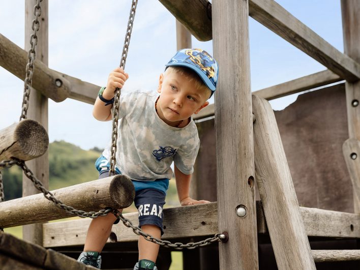 Kleiner Junge mit Kappe klettert über eine Hängebrücke aus Holzstämmen und Ketten auf den Kletterturm im Freien.
