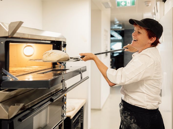 Bäckerin schiebt lachend einen Brotlaib mit dem Schieber in den Steinbackofen der Hotelbackstube.