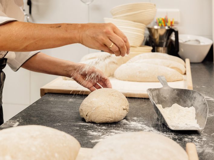 Hände bestäuben geformte Brotlaibe mit Mehl, daneben Gärkörbchen und eine Schaufel mit Mehl auf der Arbeitsfläche.