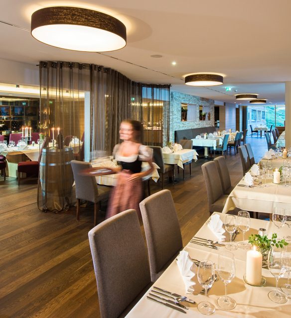 Hotelrestaurant im Almhotel Hochhäderich mit festlich eingedeckten Tischen, Kerzenlicht und großen Fenstern, eine Mitarbeiterin im Dirndl bringt lächelnd ein Tablett an den Tisch.
