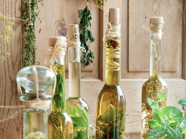 Fünf Glasflaschen mit Kräuterölen und Essig stehen vor einer Holzwand, dahinter hängen getrocknete Kräuterbüschel.