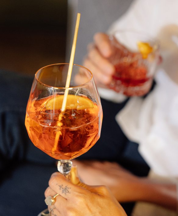 Nahaufnahme eines orangefarbenen Aperitifs mit Orangenscheibe, dahinter hält jemand einen roten Drink.