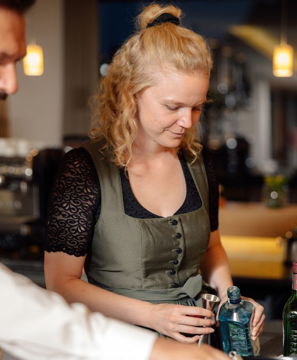 Mitarbeiterin im grünen Dirndl mixt hinter der Theke einen Drink und misst Gin mit dem Barmaß ab.