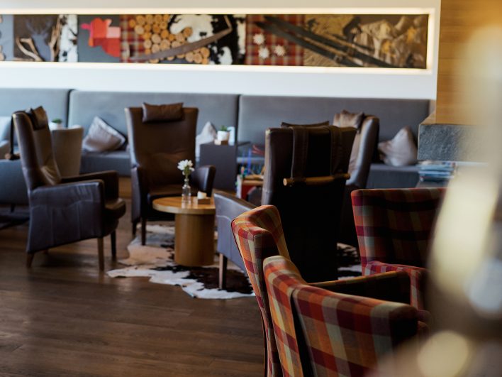 Gemütliche Sitzgruppen in der Wälderbar mit Ledersesseln, karierten Ohrensesseln und Kuhfellteppich, darüber ein Wandbild mit alpinen Motiven wie Holzscheiten, Edelweiß und alten Holzskiern.