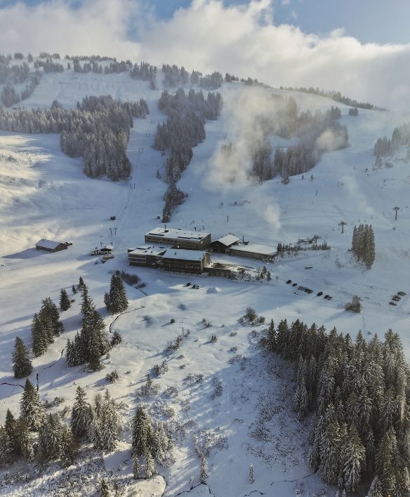 Luftaufnahme des tief verschneiten Almhotels Hochhäderich am Fuß der Pisten der Alpenarena, umgeben von Fichtenwald und aufsteigendem Nebel.