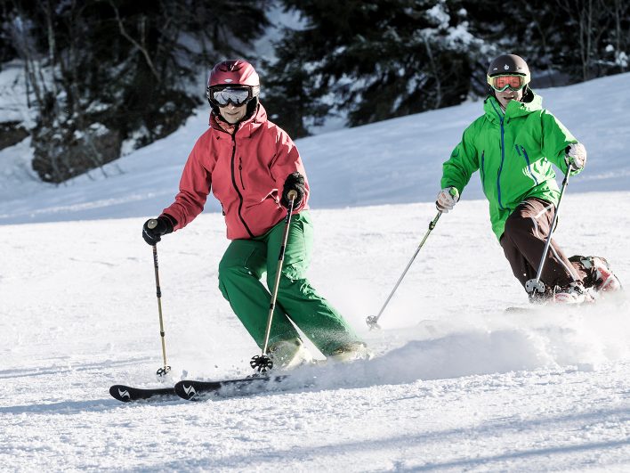 Zwei Skifahrer in roter und grüner Jacke fahren nebeneinander über die sonnige Piste der Alpenarena Hochhäderich, vor dunklen Fichten stiebt der Schnee.