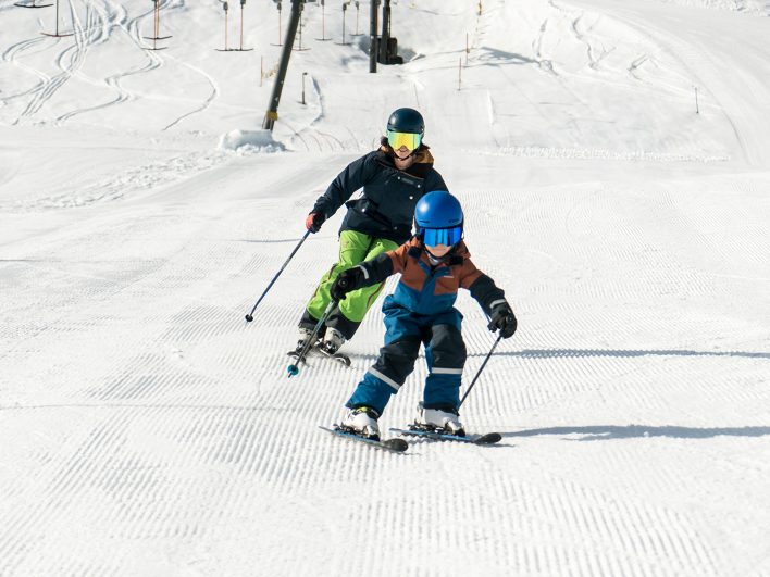 Kind im blauen Helm übt seine ersten Schwünge auf dem sanften Anfängerhang, dicht gefolgt von einer erwachsenen Begleitung, dahinter der Babylift.
