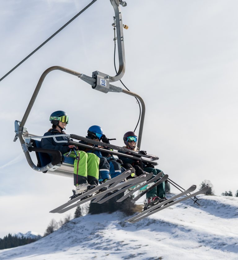 Drei lachende Skifahrer mit Helm und bunter Skibrille fahren im kindergesicherten Vierersessellift der Alpenarena Hochhäderich bergauf.