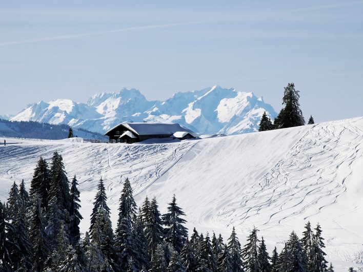 Tief verschneite Hennenmoos-Alm auf einer Kuppe im Skigebiet Alpenarena Hochhäderich, umgeben von Fichten und Skispuren, dahinter leuchtende Schneegipfel.