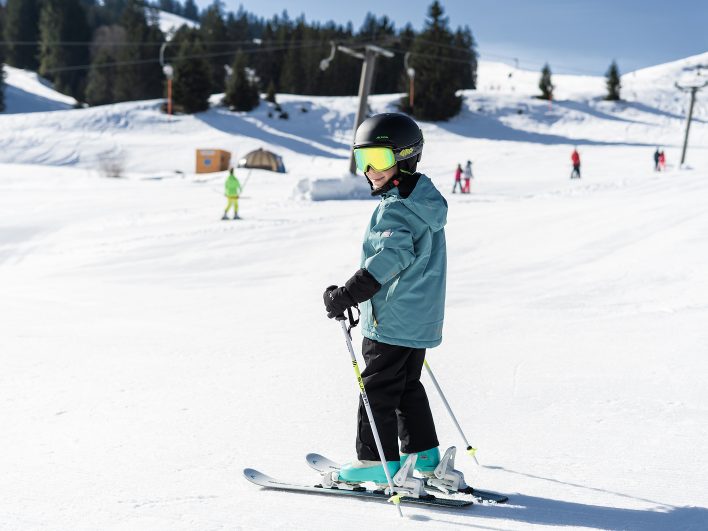 Kind mit Skibrille und Stöcken steht auf der präparierten Piste am Babylift und blickt fröhlich über die Schulter.