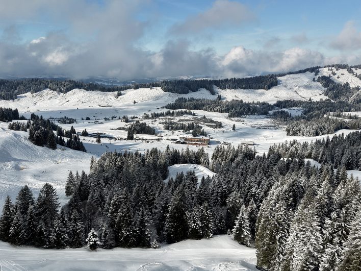 Luftaufnahme des Almhotels Hochhäderich auf 1.300 Metern, eingebettet zwischen verschneiten Fichtenwäldern, Pisten und der weiten Hochebene im Bregenzerwald.