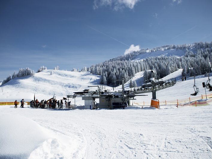Talstation des Vierersessellifts der Alpenarena Hochhäderich mit wartenden Skifahrern, dahinter frisch verschneite Pisten und Fichtenwälder unter blauem Himmel.