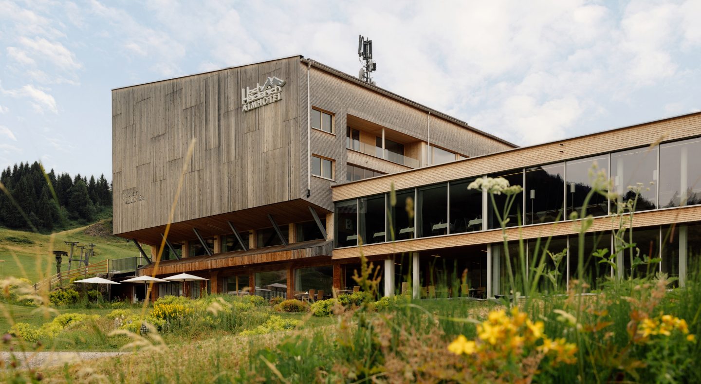 Almhotel Hochhäderich im Sommer: Holzfassade mit Schriftzug und Panoramafenstern, davor eine blühende Almwiese.