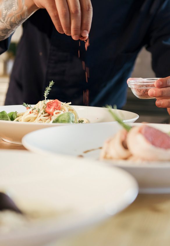 Koch streut Gewürze über einen angerichteten Teller mit Nudeln, Tomate und Thymian, daneben weitere Teller.