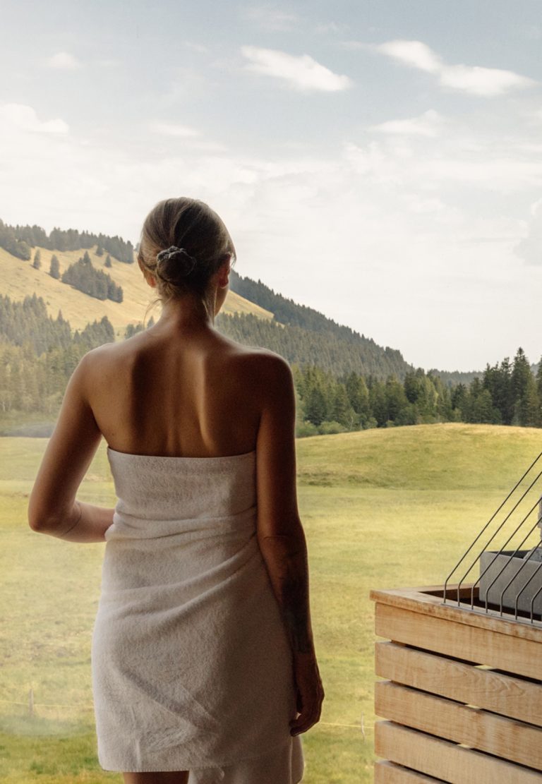 Frau im Saunatuch blickt durch das Panoramafenster der Sauna auf Almwiesen und bewaldete Hügel im Sommer.