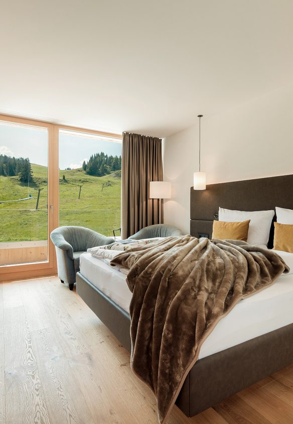 Hotelzimmer mit Doppelbett und Fellplaid, bodentiefes Fenster mit Blick auf die grüne Almwiese und den Skilift.