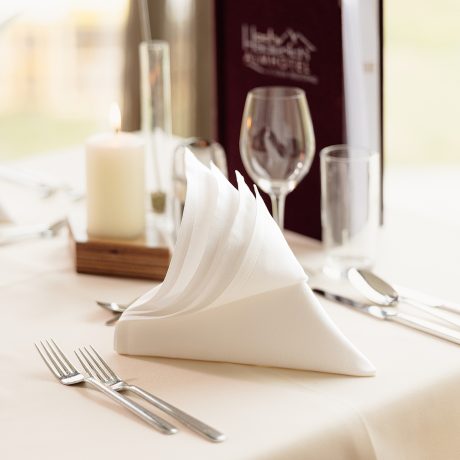 Gedeckter Tisch im Restaurant mit gefalteter Stoffserviette, Weinglas, Kerze und Getränkekarte mit Hotellogo.