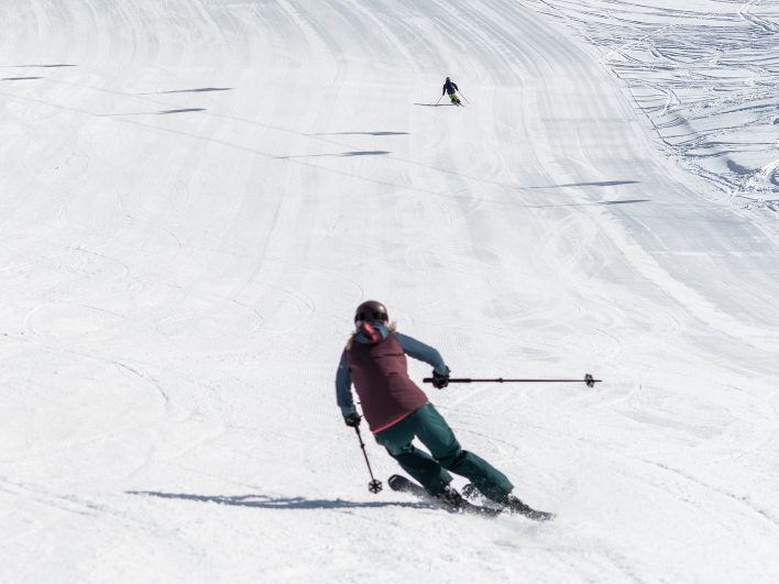 Skifahrerin zieht einen sauberen Carvingschwung auf der breiten, frisch präparierten Piste, weiter unten fährt ein zweiter Skifahrer ab.
