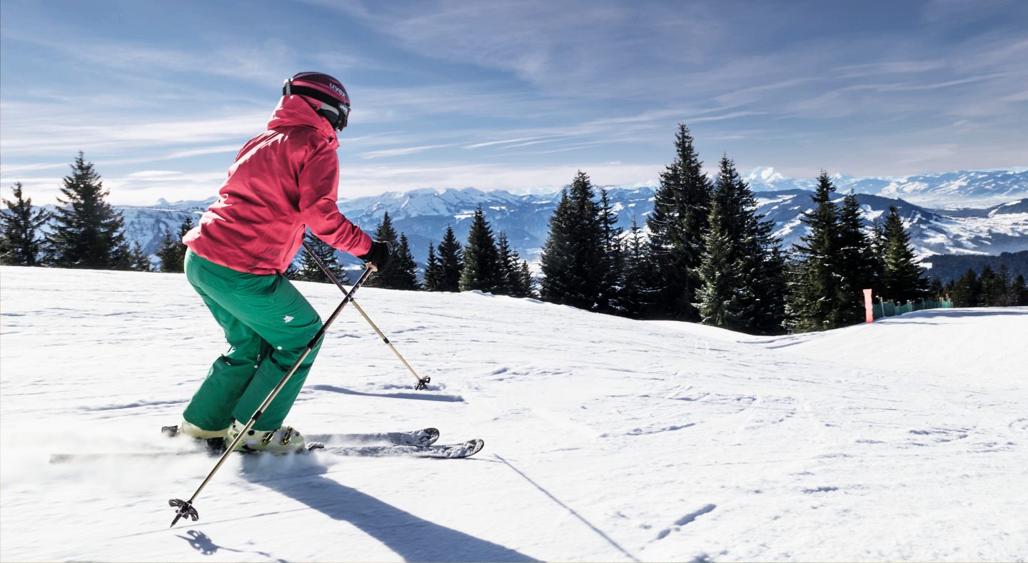 Skifahrerin in roter Jacke fährt über eine sonnige Piste, dahinter reicht der Blick über Fichten bis zu den verschneiten Bergketten der Alpen.