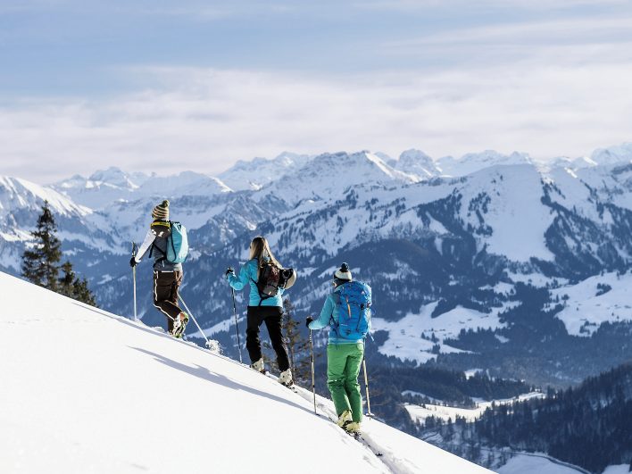 Drei Skitourengeherinnen steigen mit Fellen durch den unberührten Tiefschnee bergauf, vor ihnen ein weites Panorama verschneiter Gipfel über dem Nebelmeer.