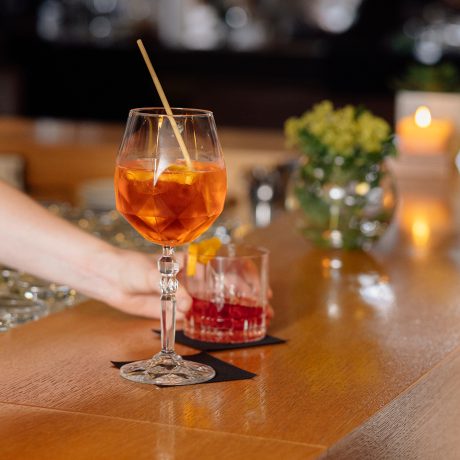 Hand stellt einen orangefarbenen Aperitif auf die Holztheke der Wälderbar, daneben ein Drink mit Orangenzeste.