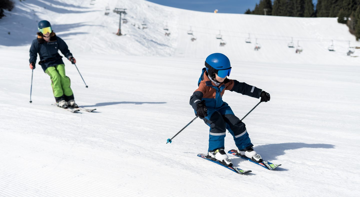 Kleiner Skifahrer in blauem Anzug zieht vor einer erwachsenen Begleitung seine Schwünge auf der präparierten Piste, dahinter der Vierersessellift der Alpenarena Hochhäderich.