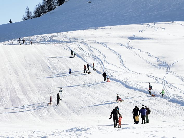 Familien mit Schlitten ziehen den sonnigen Rodelhang am Almhotel Hochhäderich hinauf, während andere Kinder schon wieder durch den Schnee hinuntersausen.